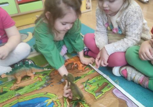 Dzieci siedzą wokół planszy edukacyjnej i ustawiają dinozaury w odpowiednich miejscach.