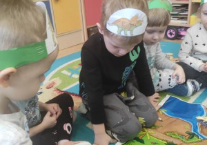 Dzieci siedzą wokół planszy edukacyjnej i ustawiają dinozaury w odpowiednich miejscach.