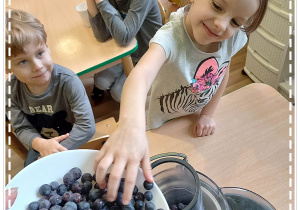 Idalia wrzuca owoce borówki do sokowirówki pod okiem nauczycielki.