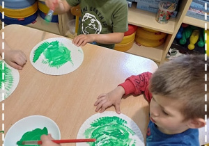 Antoś i Olek malują zieloną farbą talerzyki papierowe