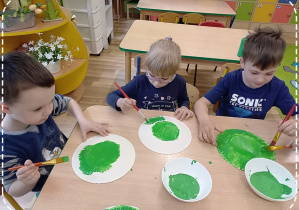 Filip M., Weronika i Tymek malują zieloną farbą talerzyki papierowe