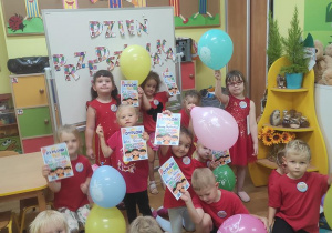 Dzieci ustawione przed tablicą z balonami, dyplomami i odznakami z okazji Dnia Przedszkolaka.