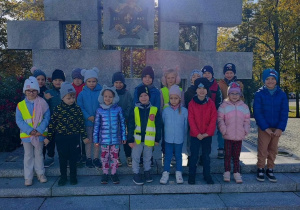 Dzieci stoją przy pomniku poległych harcerzy i harcerek.