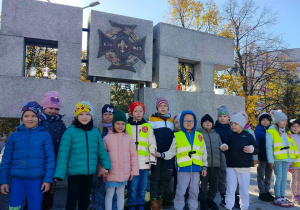 Dzieci stoją przy pomniku poległych harcerzy i harcerek.
