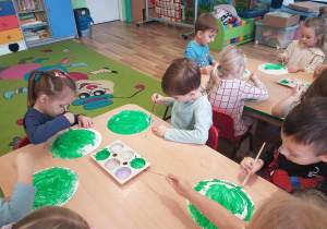 Dzieci malują papierowe talerzyki zieloną farbą.