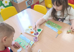 Dzieci malują kwiaty na łące i mocują na wykałaczkach owady.